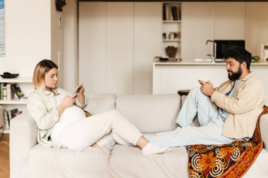 Sakallı adam ve hamile kadın evdeki koltukta otururken cep telefonu kullanıyorlar.