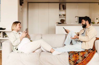 Sakallı adam ve hamile kadın evdeki koltukta otururken cep telefonu kullanıyorlar.
