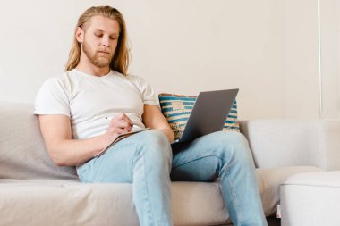 Evde kanepede otururken dizüstü bilgisayarla çalışan sakallı sarışın adam.