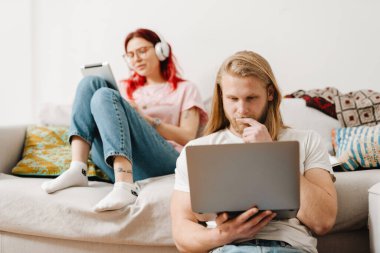 Beyaz adam ve kadın evdeki koltukta ve yerde otururken alet kullanıyorlar.