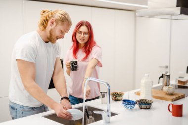 Beyaz adam evde bulaşıkları yıkıyor ve kız arkadaşıyla konuşuyor.