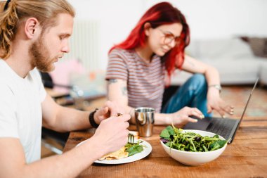 Genç beyaz çift dizüstü bilgisayar kullanıyor ve masada otururken yemek yiyorlar.