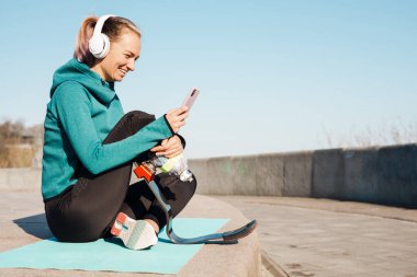 Kulaklıklı, protezli genç bir sporcu dışarıda cep telefonu kullanarak spor minderinin üzerinde oturuyor.