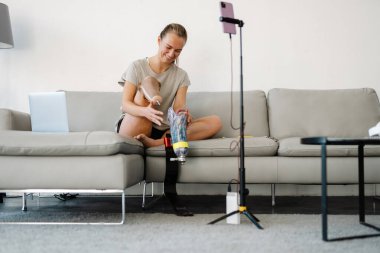 Gülümseyen beyaz engelli kadın blogcu protez bacaklı bir kadın kanepenin önünde oturmuş evde video çekiyor.