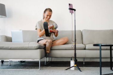 Gülümseyen beyaz engelli kadın blogcu protez bacaklı bir kadın kanepenin önünde oturmuş evde video çekiyor.