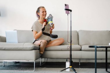 Gülümseyen beyaz engelli kadın blogcu protez bacaklı bir kadın kanepenin önünde oturmuş evde video çekiyor.