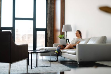 Gülümseyen, sakat, protez bacaklı bir kadın evinde dizüstü bilgisayarla koltukta oturuyor.