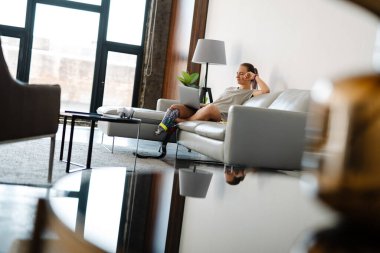 Gülümseyen, sakat, protez bacaklı bir kadın evinde dizüstü bilgisayarla koltukta oturuyor.