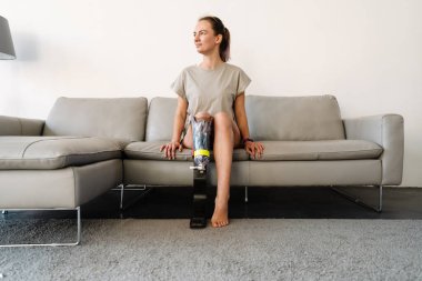 Gülümseyen, sakat, protez bacaklı bir kadın evdeki kanepede oturuyor.