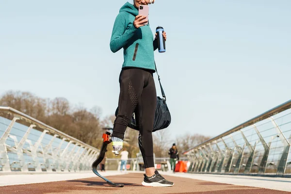 一个年轻的健康的女运动员站在那里 拿着手机和瓶子里的水 站在室外喝着瓶子里的形象 — 图库照片