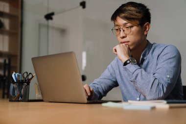 Ofiste otururken dizüstü bilgisayarla çalışan gözlüklü Asyalı adam.