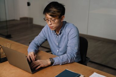 Ofiste otururken dizüstü bilgisayarla çalışan gözlüklü Asyalı adam.