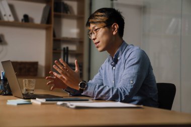 Ofiste dizüstü bilgisayarla video görüşmesi yaparken gözlüklü memnun Asyalı adam el kol hareketi yapıyor.