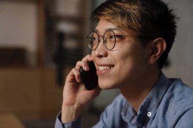 Ofiste çalışırken cep telefonuyla konuşan gözlüklü mutlu Asyalı adam.