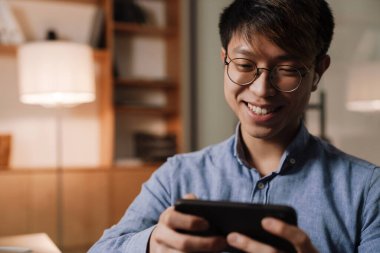 Kulaklıklı gülümseyen Asyalı adam ofiste cep telefonuyla video oyunu oynuyor.