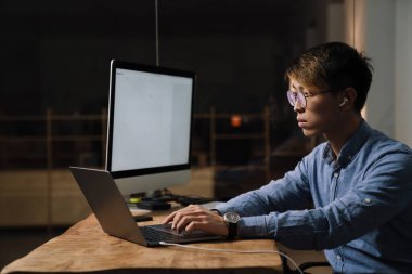 Kulaklıklı Asyalı adam bilgisayarda ve laptopta çalışıyor.