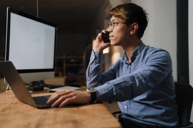 Ofisteki bilgisayarlarla uğraşırken cep telefonuyla konuşan Asyalı adam.