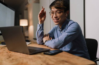 Kulaklıklı gülümseyen Asyalı adam ofiste dizüstü bilgisayarla konuşurken el kol hareketi yapıyor.