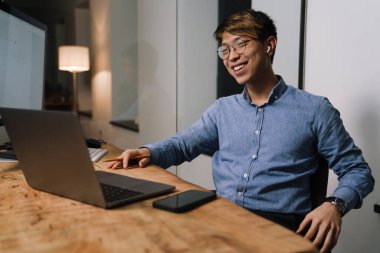 Ofiste dizüstü bilgisayarla çalışırken gülümseyen kulaklıklı mutlu Asyalı adam.