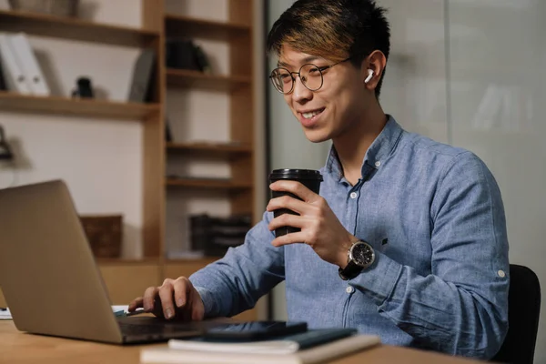 オフィスでラップトップで作業しながら イヤホンコーヒーを飲みながらアジアの男を笑顔 — ストック写真