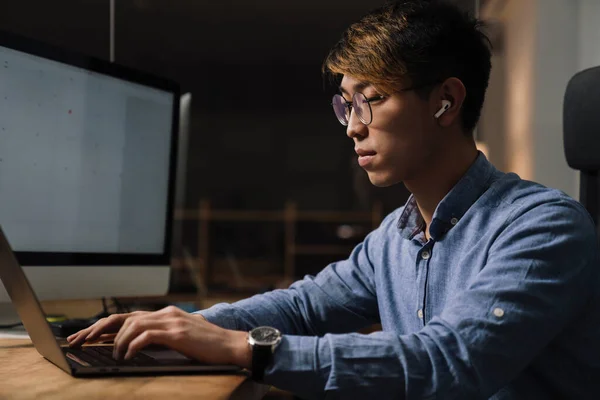 Keskittynyt Aasialainen Kaveri Kuulokkeissa Työskentelee Kannettavan Tietokoneen Kanssa Istuessaan Pöydässä — kuvapankkivalokuva
