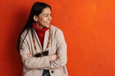 Palto giymiş genç İspanyol kadın gülümsüyor ve turuncu duvarın ötesine bakıyor.