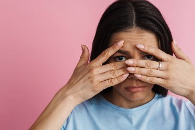 Genç, korkmuş bir kadın yüzünü kapatıp pembe duvarın üzerinden izole edilmiş kameraya bakıyor.