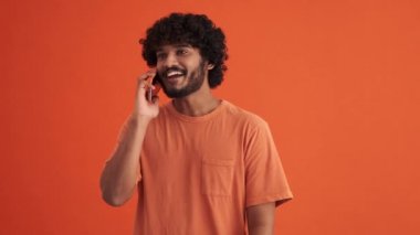 Turuncu stüdyoda gülümseyen kıvırcık saçlı Hintli adam telefonla konuşuyor.