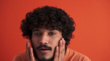Pozitif kıvırcık saçlı Hintli adam turuncu stüdyodaki kameraya bakışını düzeltiyor.