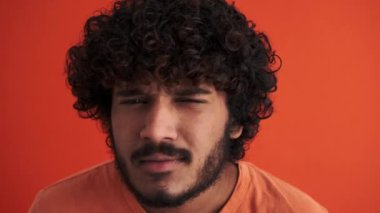 Ciddi kıvırcık saçlı Hintli adam turuncu stüdyoda kameraya bakıyor.