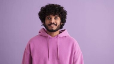 Neşeli kıvırcık saçlı Hintli adam mor stüdyoda kameraya el sallıyor.