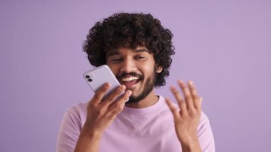 Kıvırcık saçlı gülümseyen Hintli adam mor stüdyoda telefonla sesli mesaj kaydediyor.