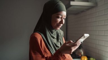 Neşeli Müslüman kadın telefonla yazıyor ve mutfaktaki pencereye bakıyor.