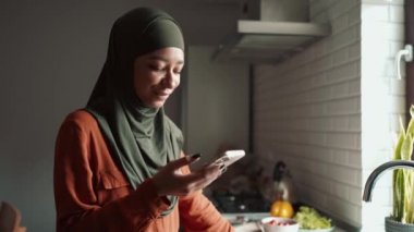 Telefonla yazan ve mutfaktaki pencereye bakan gülen Müslüman kadın.