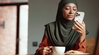 Gülümseyen Müslüman kadın evdeki masada bir fincan çayla telefonla mesaj atıyor.