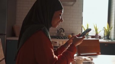Neşeli Müslüman kadın evde telefonla mesaj atıyor.
