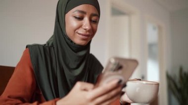 Genç Müslüman kadın çay içiyor ve evde telefonla yazıyor.