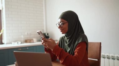 Gözlüklü neşeli Müslüman kadın evde telefonla mesaj atıyor.
