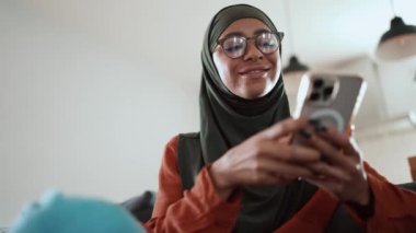 Mutlu Müslüman kadın gözlük takıyor. Kanepede telefonla mesajlaşıyor.