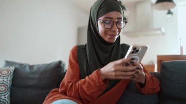 Kanepede telefonla mesajlaşan gözlüklü, pozitif Müslüman bir kadın.