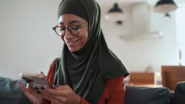 Gözlüklü, gülümseyen Müslüman kadın kanepede telefonla mesajlaşıyor.