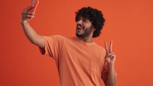 陽気な巻き毛のインド人男作る自画によって電話とショー異なるジェスチャーでザオレンジ色のスタジオ — ストック動画