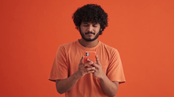 Fröhlich Gelockter Indischer Mann Schaut Orangefarbenen Studio Auf Das Telefon — Stockvideo