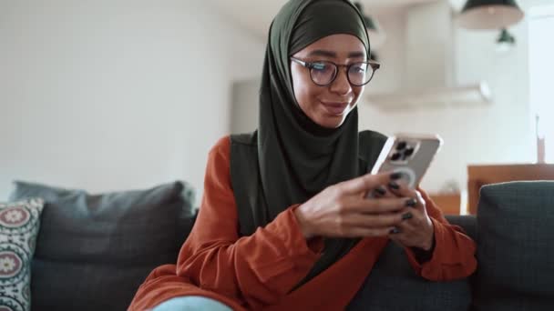 积极的穆斯林妇女戴着眼镜 在沙发上通过电话发短信 — 图库视频影像