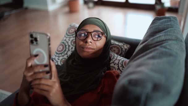 眼鏡をかけたイスラム教徒の女性がソファの上で電話でテキストメッセージ — ストック動画