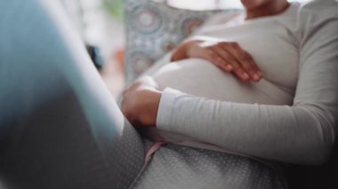 Afrikalı hamile kadın evdeki kanepede karnını okşuyor.