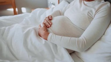 Afrikalı hamile bir kadın evdeki yatakta karnını okşuyor.