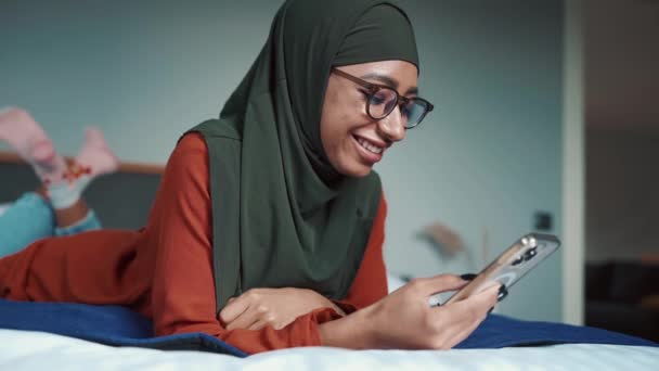 在家里 戴着眼镜在床上用电话打字的穆斯林妇女笑着 — 图库视频影像