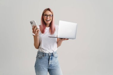 Beyaz arka planda izole edilmiş dizüstü bilgisayar ve cep telefonuyla poz verirken gülümseyen genç kadın