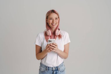 Kulaklıklı, gülümseyen ve cep telefonu kullanan genç Avrupalı kadın beyaz arka planda izole edilmiş.